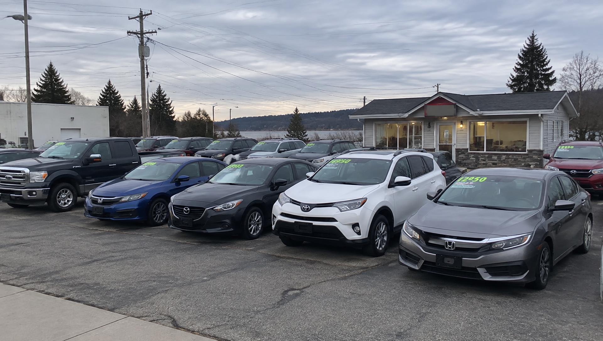 Small independent used-car lot with priced vehicles parked outside a sales office